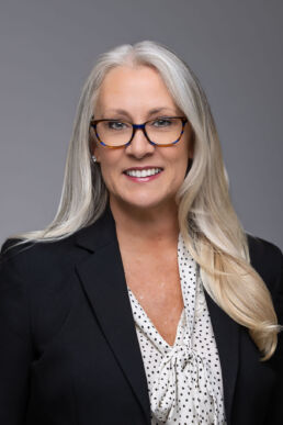 Corporate headshot of female executive with gray hair and glasses against a dark gray background.