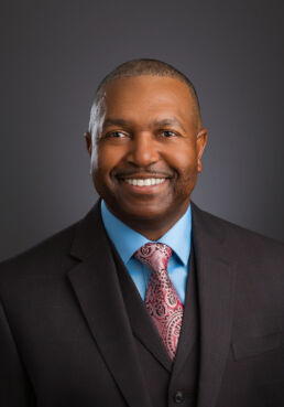 Professional business portrait in studio setting of older man wearing a dark suit with a blue shirt and pink tie.