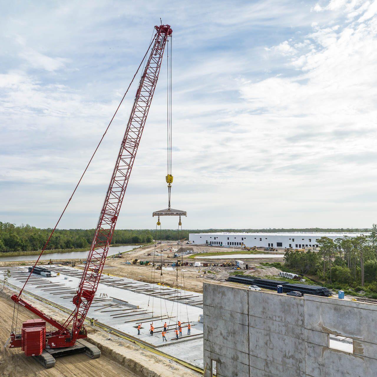 Aerial drone photograph of a construction site in Tampa, Florida showing a red crane lifting a concrete wall panel with workers guiding the placement.