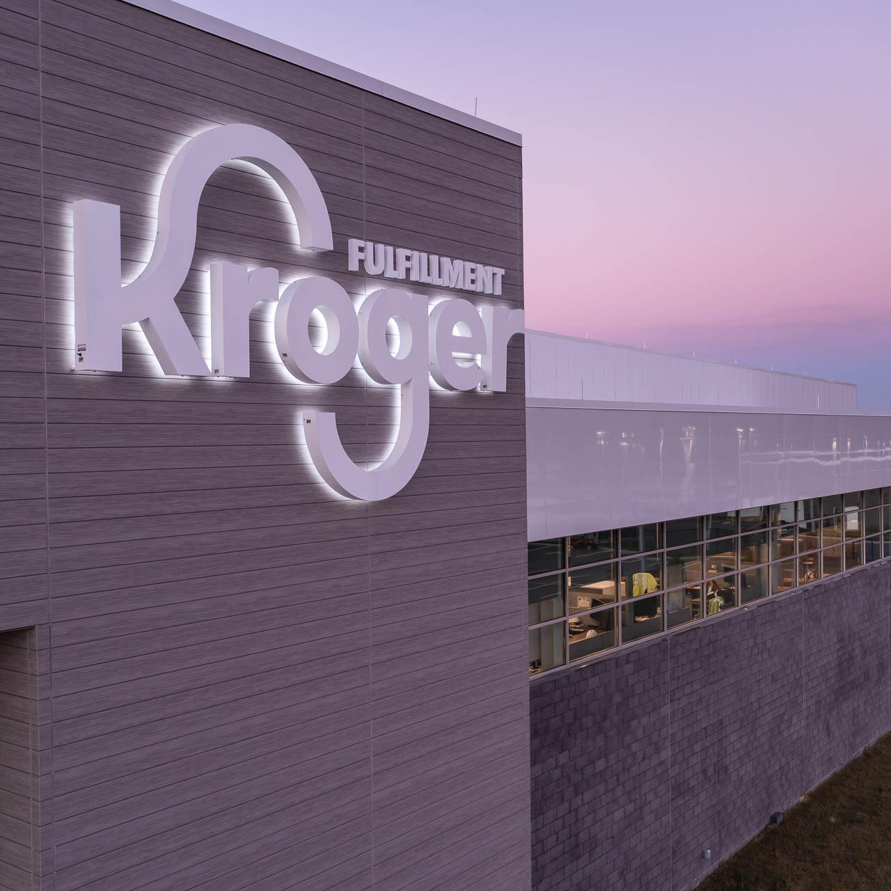 Aerial drone photograph of the Kroger fulfillment center exterior at dusk with illuminated signage and modern architectural detail.