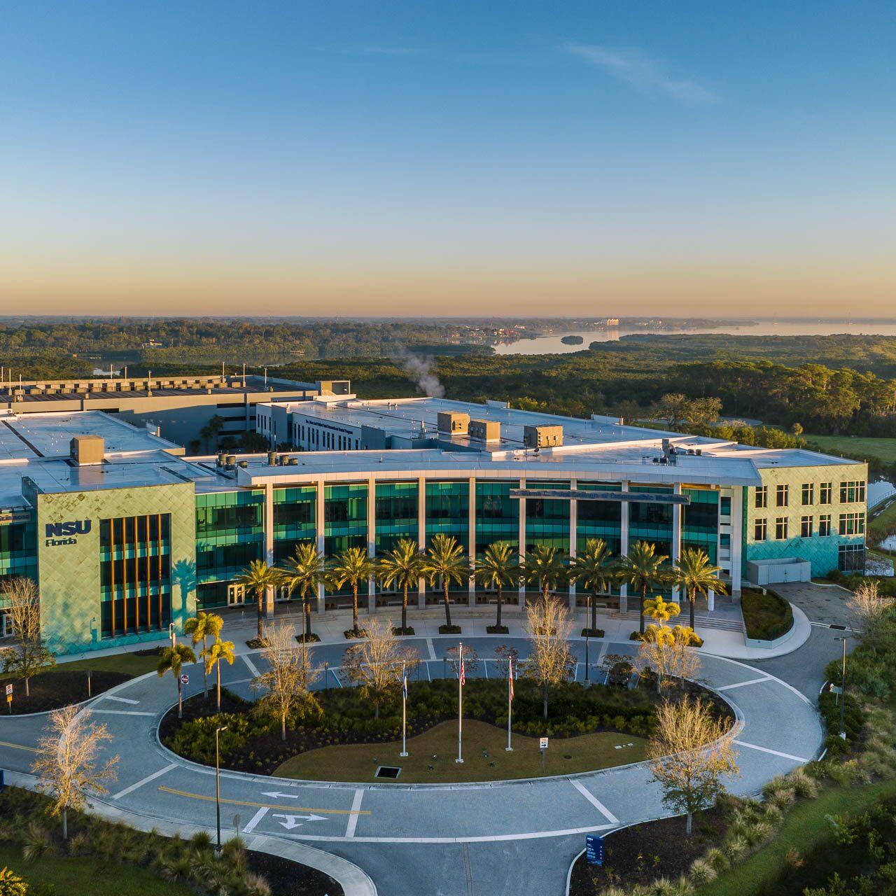 Aerial drone photograph of Nova Southeastern University in Clearwater, Florida showing the modern glass-front campus building surrounded by water and greenery.