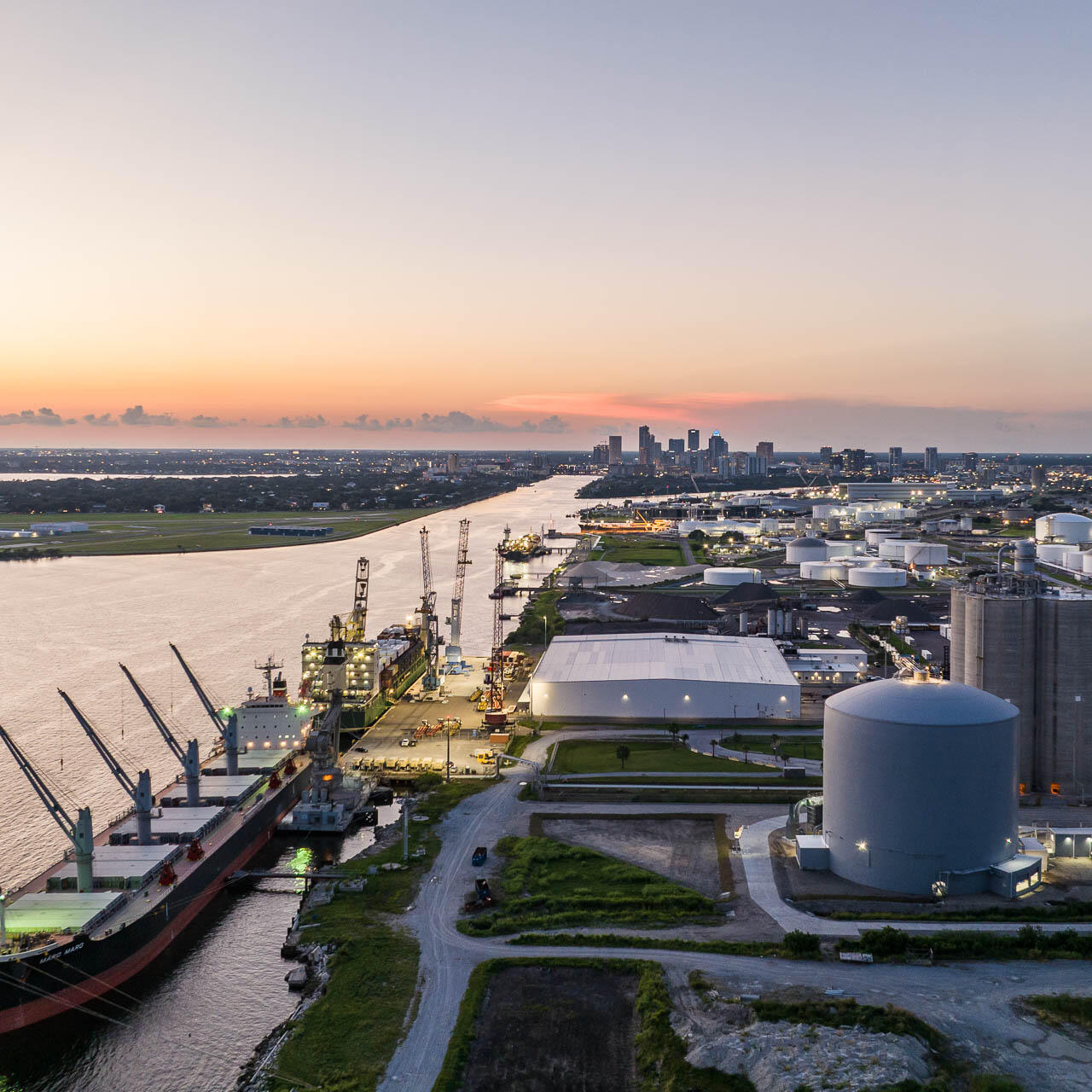 Aerial drone photograph of the Port of Tampa at dusk showing cargo ships, industrial storage tanks, and downtown skyline in the distance.